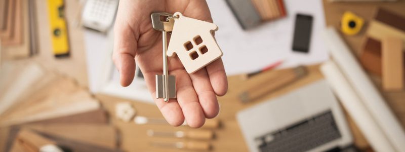 39380580_s 39380580 - real estate agent handing over a house key, desktop with tools, wood swatches and computer